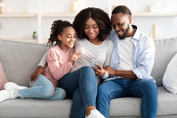 Família afro-americana feliz segurando e usando celular em casa — Fotografia de Stock