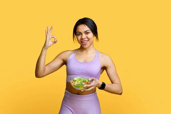 Ajuste joven mujer negra recomendando una nutrición saludable, sosteniendo un tazón con ensalada de verduras y mostrando un gesto aceptable —  Fotos de Stock