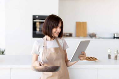 Mutfakta yemek pişiren, önlük giyen ve dijital tabletle yemek tarifleri inceleyen, elinde kızartma tavası tutan mutlu bir ev hanımı.