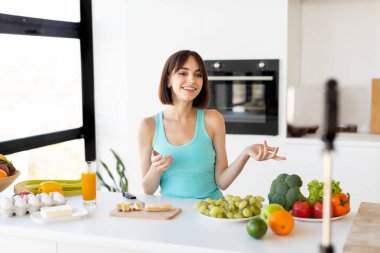 Yemek blogcusu kavramı. Milenyum kadını akıllı telefondan yeni video tarifi kaydediyor, modern mutfağın içinde duruyor.