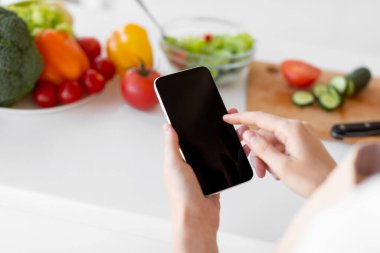 Sağlıklı gıda ve diyet planlama konsepti. Genç bir kadın boş ekranlı akıllı telefon kullanıyor, mutfakta salata hazırlıyor.