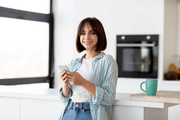Glimlachende jonge dame met behulp van smartphone, terwijl ze thuis geniet van koffie in de keuken, bij de tafel staat en lacht naar de camera — Stockfoto