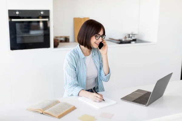 Práce z domova. Freelancer pomocí notebooku, mluvení na smartphone a dělat si poznámky, sedí v kuchyni interiéru — Stock fotografie