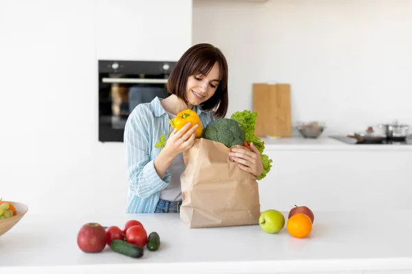 Тысячелетняя женщина распаковывает бумажный пакет со свежими органическими овощами и фруктами, стоя на современной кухне — стоковое фото
