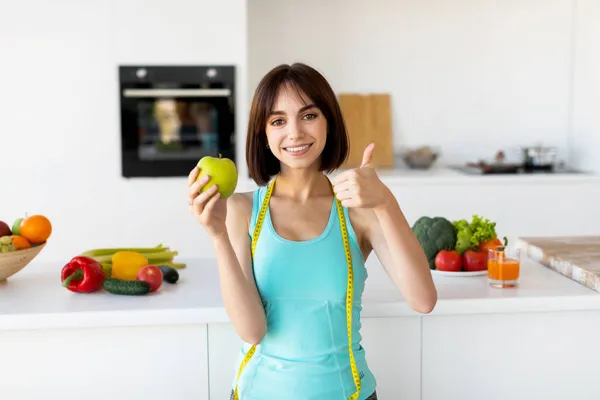 Joven mujer delgada sosteniendo manzana y mostrando el pulgar hacia arriba, recomendando una alimentación saludable, posando en el interior de la cocina — Foto de Stock