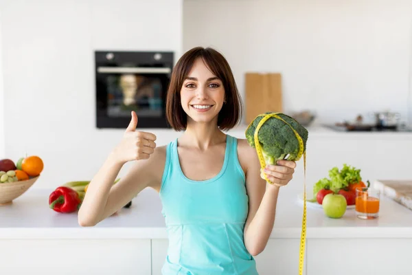 Концепция диеты. Счастливая стройная женщина держит брокколи с измерительной лентой и показывает большой палец вверх, кухонный интерьер — стоковое фото
