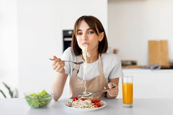 Glad dam äter spagetti och grönsakssallad, njuter av god lunch, sitter vid bordet och tittar på kameran i köket — Stockfoto