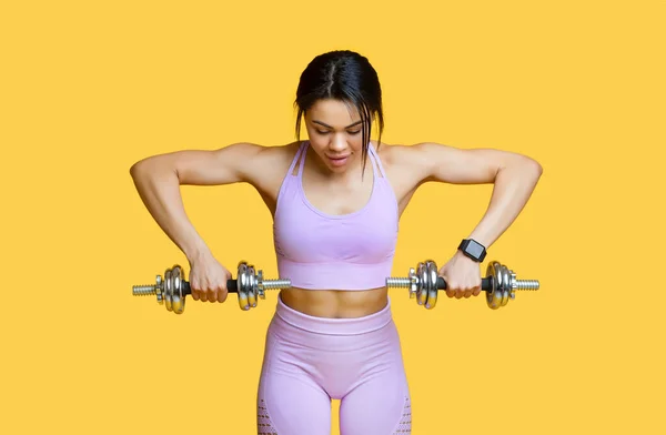Concepto deportivo. Mujer afroamericana en forma joven haciendo ejercicio con dos mancuernas, haciendo ejercicios de brazos, fondo amarillo — Foto de Stock