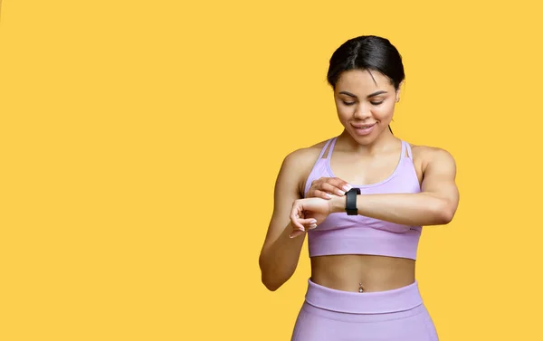 Glad afrikansk amerikansk dam använder smartwatch för att övervaka utvecklingen, kontrollera hennes prestanda, gul bakgrund — Stockfoto