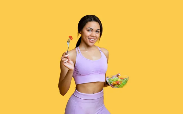 Dieta e perdita di peso. Felice signora afro-americana mangiare insalata di verdure fresche, sorridente alla macchina fotografica, sfondo giallo — Foto Stock