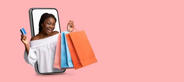 Spännande ung svart dam som visar kreditkort och presentpåsar, kommer ut ur mobiltelefon skärm på rosa bakgrund — Stockfoto