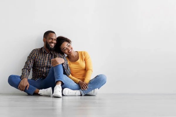 Soddisfatto giovane afroamericano maschio e femmina seduto sul pavimento su sfondo grigio muro, abbracci, pianificazione interna — Foto Stock