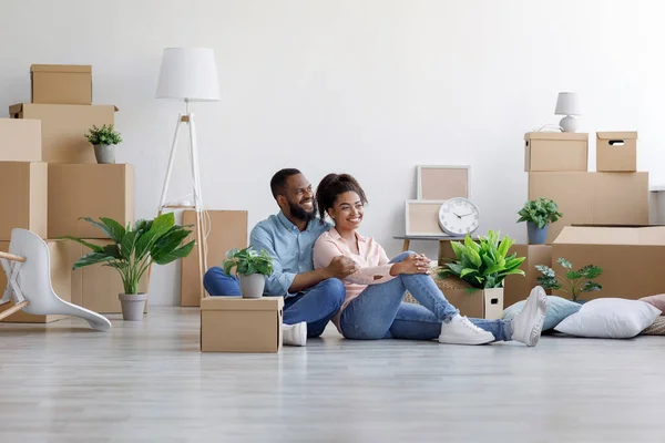 Alegre joven marido negro y esposa descanso, sentarse en el suelo entre cajas de cartón con cosas y plantas —  Fotos de Stock