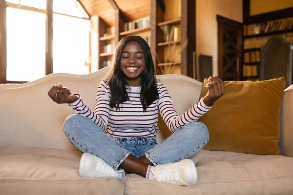 Gestión del estrés, bienestar, atención plena. Calma joven dama negra meditando con los ojos cerrados en el sofá en casa —  Fotos de Stock