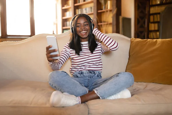 Fröhliche schwarze Frau sitzt mit Kopfhörern auf der Couch, bedient sich einer neuen Musik-App auf dem Smartphone und hört zu Hause Hörbücher — Stockfoto