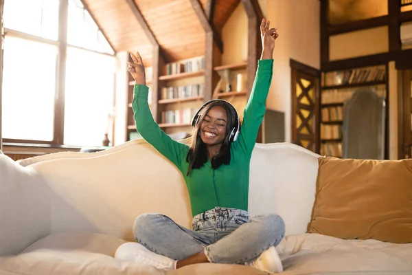 Mujer negra despreocupada escuchando música, usando auriculares, bailando en el sofá en la sala de estar, espacio libre — Foto de Stock