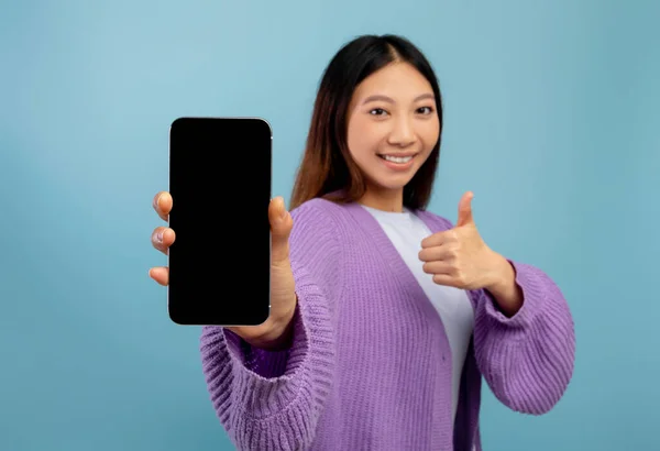 Ich empfehle. Glückliche asiatische Dame zeigt Smartphone mit leerem Bildschirm, gestikuliert mit dem Daumen nach oben und lächelt, blauer Hintergrund — Stockfoto