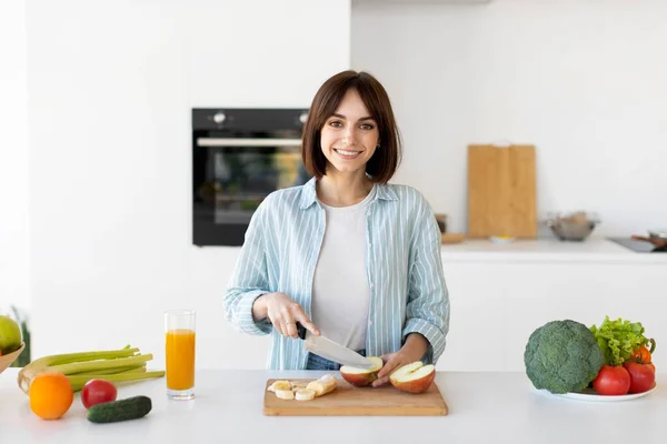 Glad ung dam skära frukt på träskiva, förbereda färsk sallad, står i köket och ler mot kameran — Stockfoto