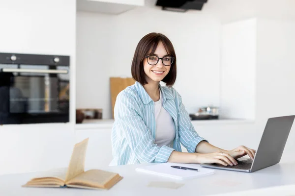Gelukkig jong freelancer werken aan laptop in keuken interieur, zitten aan tafel en glimlachen op de camera — Stockfoto