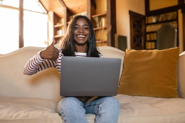 Smiling young black woman with laptop showing thumb up gesture at home, recommending online work during covid pandemic