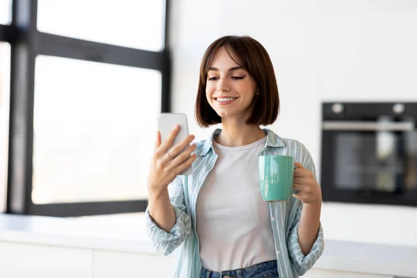 Feliz mujer milenaria utilizando el teléfono inteligente y beber té en el interior de la cocina, navegar por Internet mientras disfruta del café —  Fotos de Stock