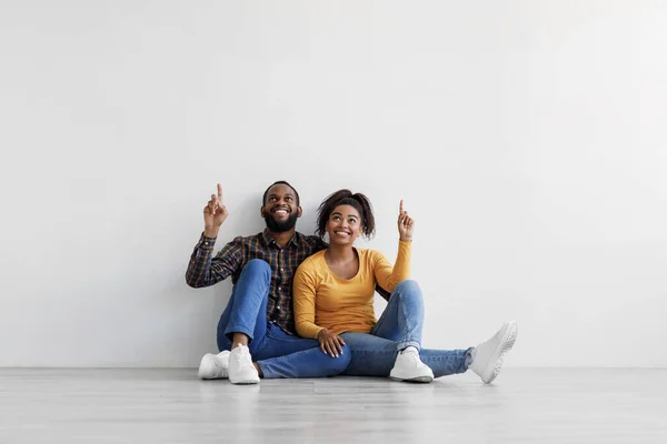 Usmívající se africká americká rodina sedí na podlaze nad šedou stěnou pozadí, ukazující prsty nahoru na kopírovací prostor — Stock fotografie