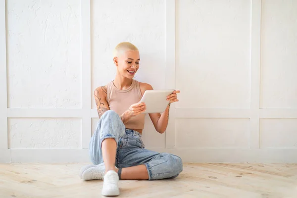 Felice giovane donna utilizzando tablet digitale, navigare in internet ot chattare con gli amici, seduto sul pavimento sopra il muro bianco — Foto Stock