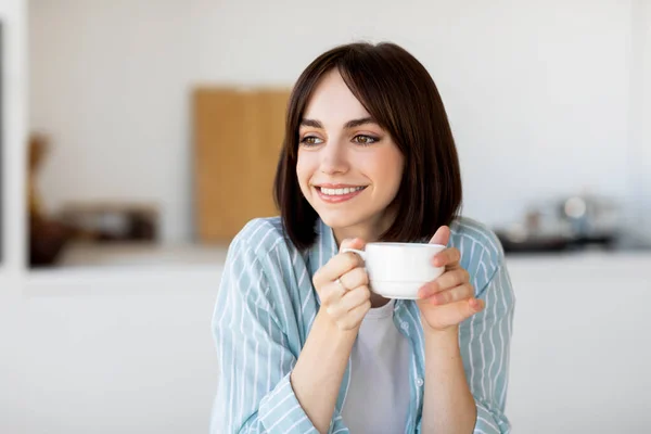 Goedemorgen koffie. Portret van jonge dame met mok in handen, zittend in keukeninterieur en opzij kijkend, vrije ruimte — Stockfoto