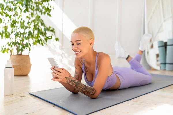 Glücklich fitte Dame, die sich auf Yogamatte ausruht, auf Smartphone im Internet surft und nach dem Fitnesstraining zu Hause eine Pause einlegt — Stockfoto