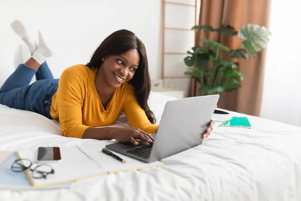 Afro-americano feminino usando laptop aprendendo online deitado no quarto — Fotografia de Stock