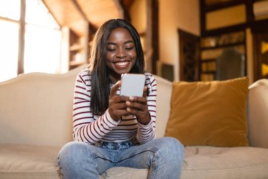 Genç siyahi kadın cep telefonu kullanıyor, sosyal medyayı kontrol ediyor, interneti arıyor, internette sohbet ediyor, evde video izliyor.