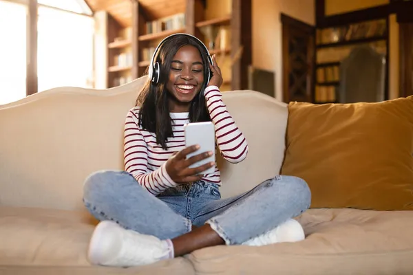 Felice giovane donna nera in cuffia ascoltare musica o audiolibro sul cellulare, seduto sul divano a casa — Foto Stock