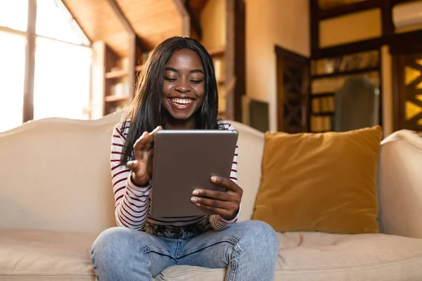Glad ung svart dam som använder surfplatta, lär sig eller arbetar på distans, har online videosamtal, sitter på soffan inomhus — Stockfoto