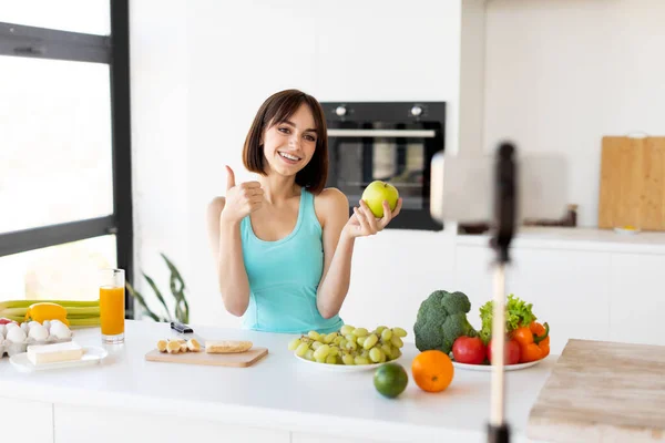 Glückliche Frau zeigt Daumen hoch und hält Apfel, Aufnahme eines neuen Videos für Sport und Food Blog, Kücheneinrichtung — Stockfoto