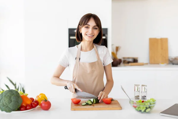 Glad ung dam matlagning färsk grönsakssallad, hugga gurka och ler mot kameran i köket — Stockfoto