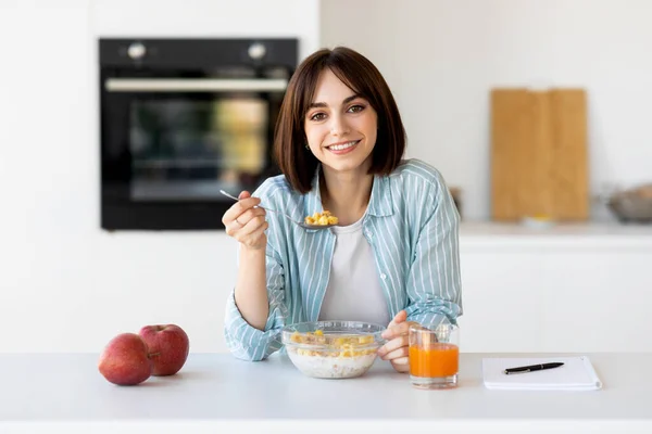 Hälsosam frukost koncept. Glad dam äter müsli skål med äpplen och juice, sitter i modernt kök interiör — Stockfoto