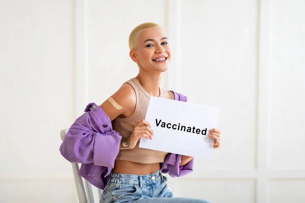 Señora emocionada con cartel vacunado sentado sobre la pared blanca, feliz mujer milenaria se vacunó contra el coronavirus — Foto de Stock