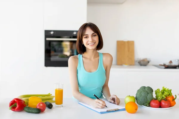 Счастливая фитнес-леди в спортивной форме писать ежедневный рацион диеты или здоровый рецепт, стоя на кухне — стоковое фото