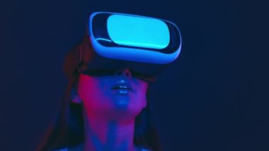 Artırılmış gerçeklik dünyası. VR kulaklıklı heyecanlı Asyalı kadın etrafa bakıyor, neon ışıklarıyla sanal video oyunu oynuyor.