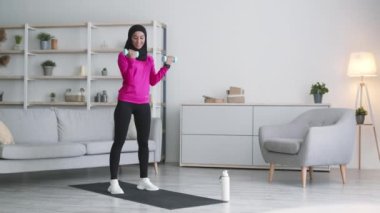 Ev antrenmanı. Geleneksel tesettürlü güçlü genç Müslüman kadın oturma odasında dambıllarla birlikte.