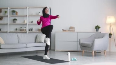 Evde dinamik fitness. Genç mutlu Müslüman kadın tesettür takıyor aktif hamleler yapıyor ve gülümsüyor.