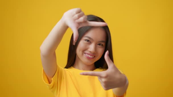 Aziatische vrouw op zoek door middel van Vinger Frame nemen van foto, gele achtergrond — Stockvideo