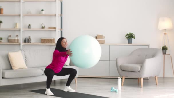 Joven mujer activa de Oriente Medio usando hijab practicando sentadillas ejercicio llevando pelota de fitness, entrenamiento en casa — Vídeos de Stock