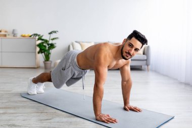 Çıplak gövdeli genç bir Arap adam, tahta üzerinde duruyor, ev içi spor yapıyor, kaslarını çalıştırıyor, evde şınav çekiyor.