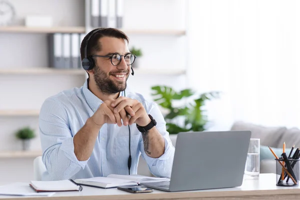 Hombre europeo adulto contento con barba en gafas, auriculares funciona en el ordenador portátil hablar con los clientes en la oficina o el interior de la habitación —  Fotos de Stock