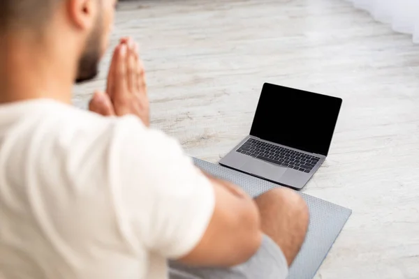 Невпізнаваний молодий арабський чоловік медитує або робить йогу перед ноутбуком з чистим екраном вдома, висміюючи — стокове фото