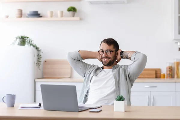 Χαρούμενος ενήλικας καυκάσιος επιχειρηματίας με γενειάδα σε γυαλιά ξεκουράζεται ενώ εργάζεται στο laptop σε μινιμαλιστικό εσωτερικό κουζίνας — Φωτογραφία Αρχείου