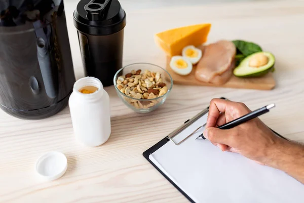 Primer plano del joven culturista escribiendo un plan de comidas en el portapapeles con maqueta, comiendo alimentos saludables, usando batidos de proteínas —  Fotos de Stock