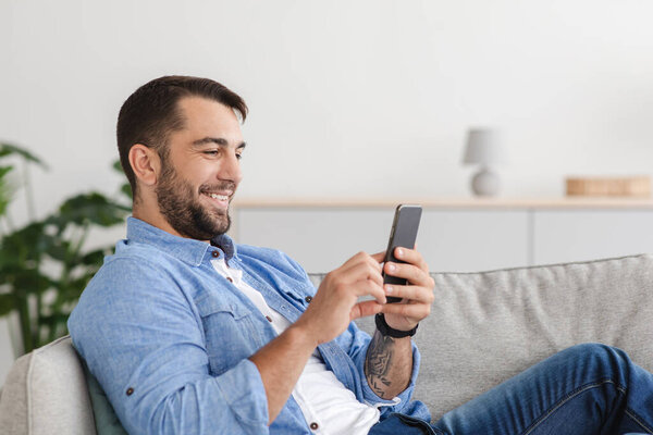 Glad mature adult caucasian man with beard sitting on sofa, chatting on smartphone