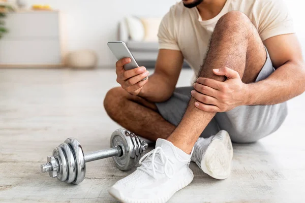 Крупный план молодого араба, сидящего на полу со смартфоном после тренировки гантели, проверка нового спортивного видео в Интернете — стоковое фото